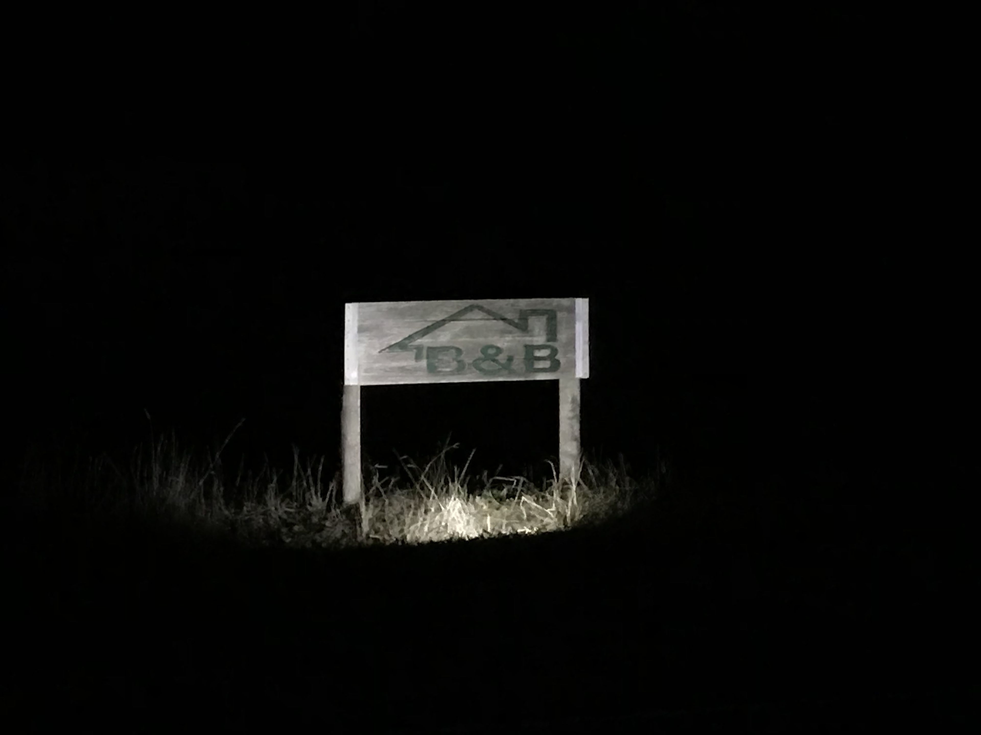 A sign with the letters "B&B" and a house symbol, set against a dark background with grass at the base.