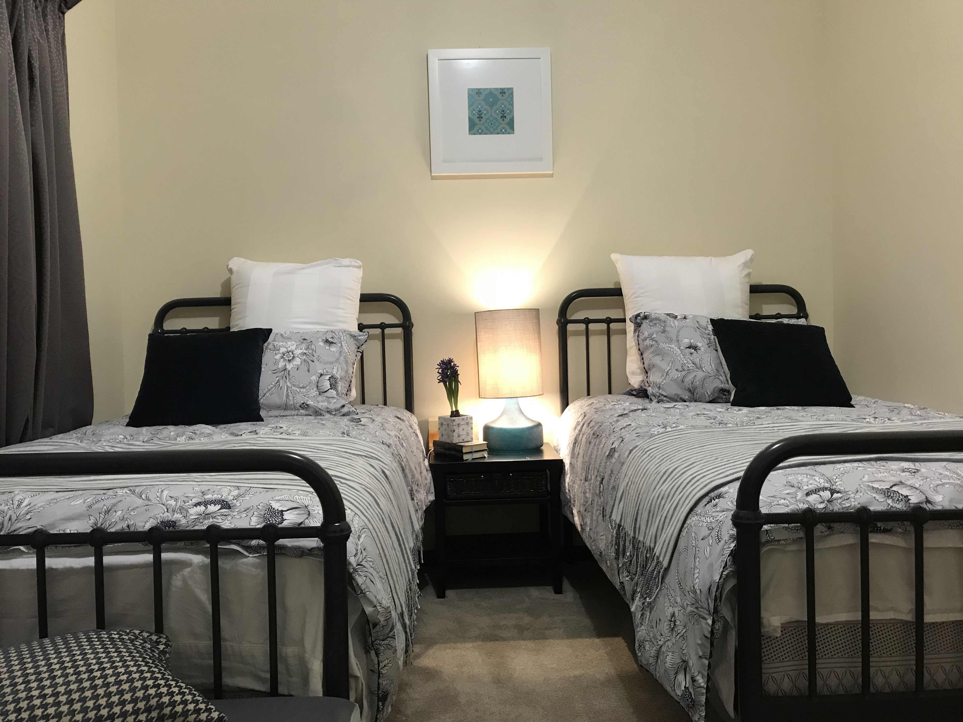 A bedroom with two single beds, each with a black metal frame and floral-patterned bedding. There is a bedside table with a lamp and a framed picture on the wall above.