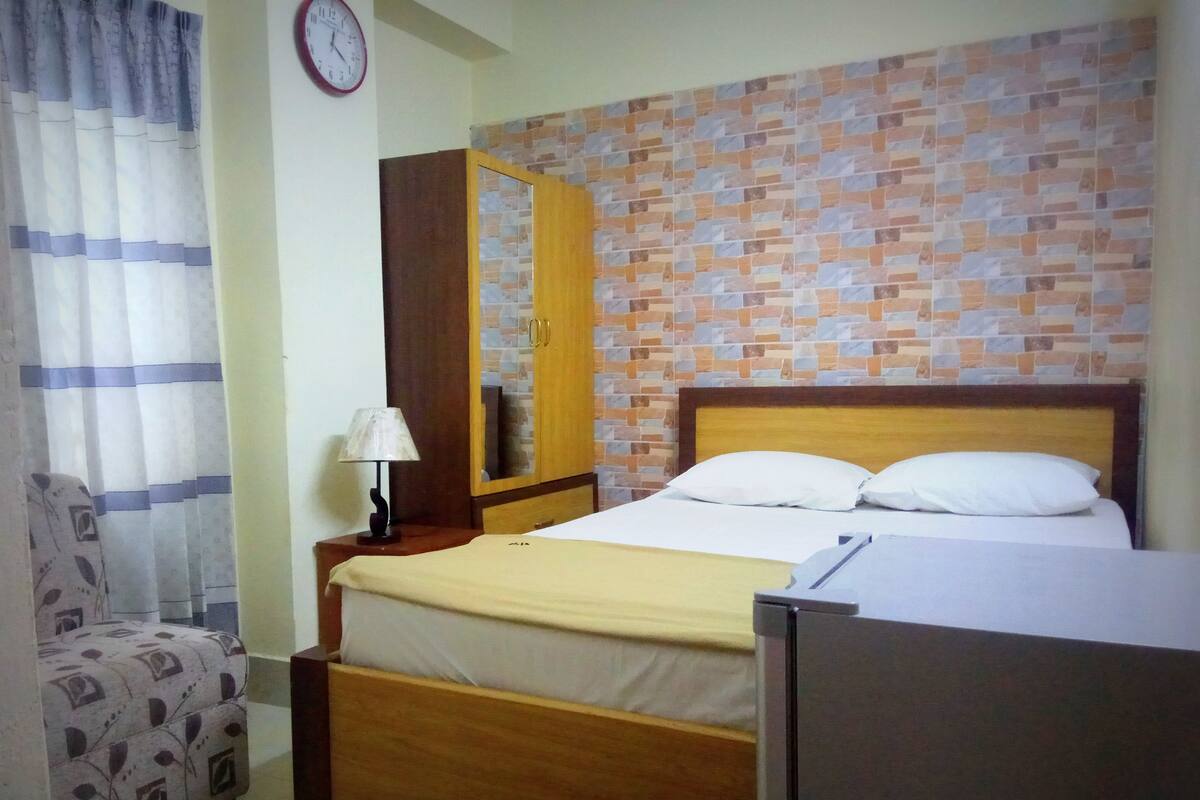 A bedroom with a wooden bed, a nightstand with a lamp, a wardrobe, a patterned armchair, and a clock on the wall.