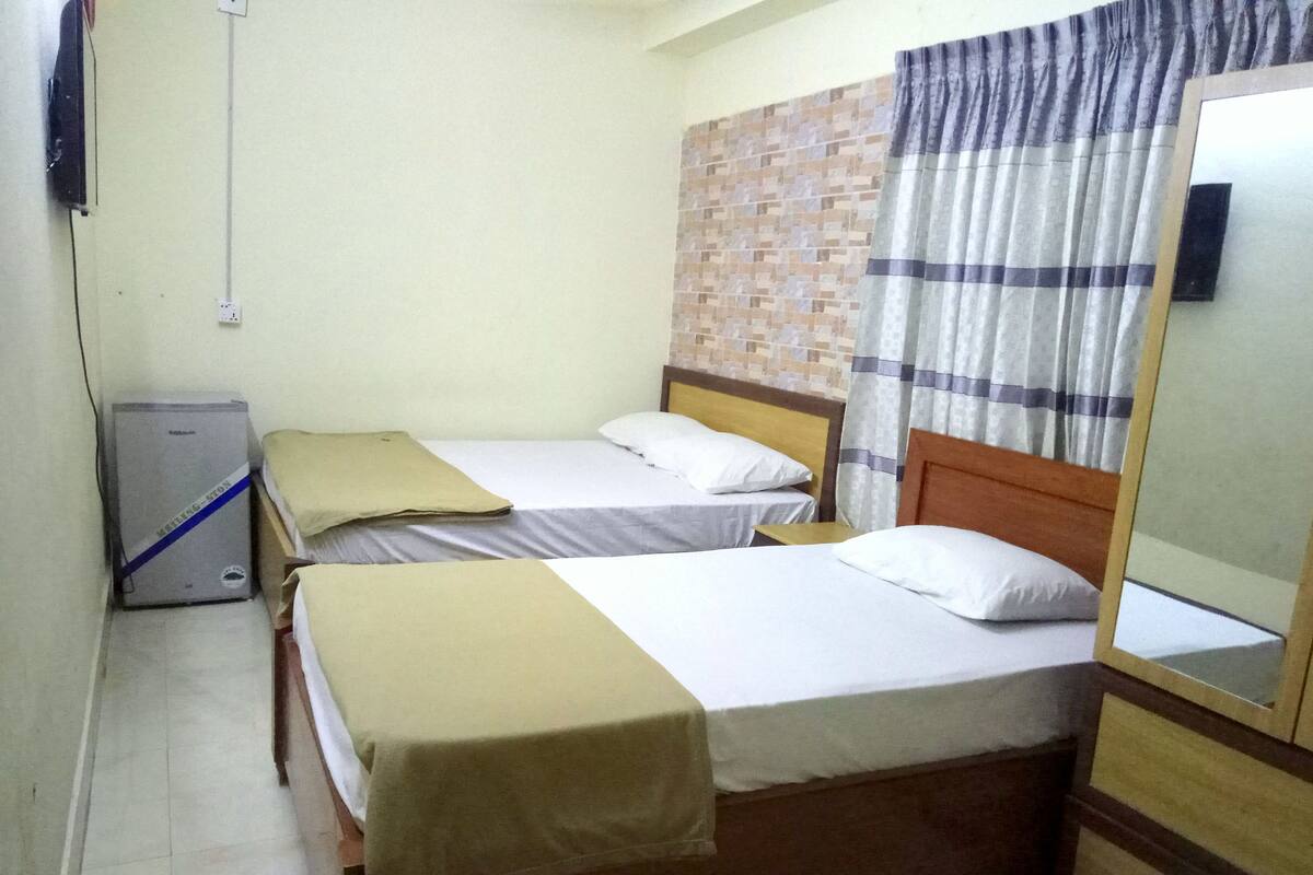 A small, simple room with two single beds, a small table, a mirror, and a wall-mounted heater.