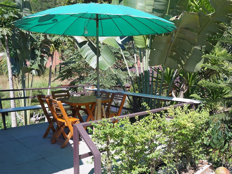Une terrasse extérieure avec une table et des chaises sous un parasol vert, entourée d’une végétation tropicale luxuriante.