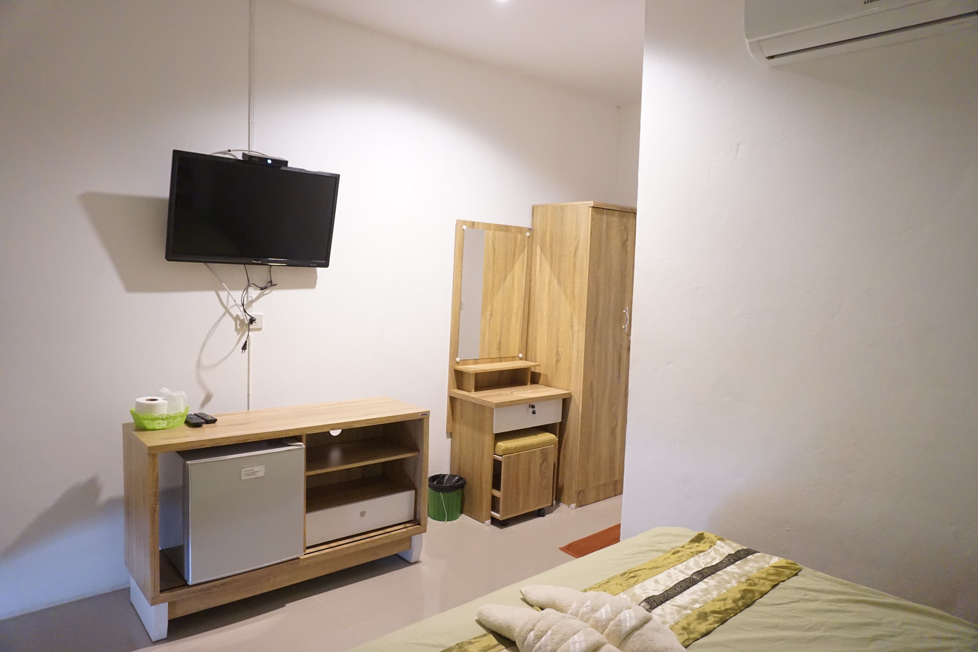 Un dormitorio con cama, televisión montada en la pared, armario de madera y un refrigerador pequeño.