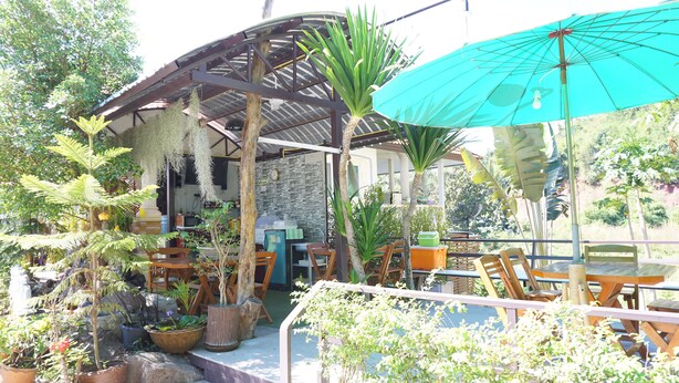 Un patio al aire libre con mesas y sillas de madera, rodeado de plantas en macetas y una zona de estar cubierta.