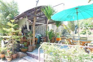 Un espace extérieur aménagé en terrasse, avec des tables et des chaises en bois, entouré de plantes en pot et doté d’un coin salon abrité.