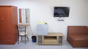 Una habitación con un armario de madera, un escritorio con una silla, un televisor montado en la pared, un refrigerador pequeño, un mueble de madera y un sofá marrón.