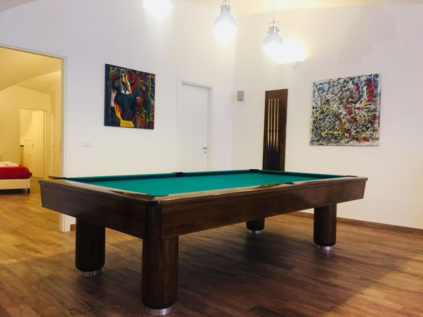 A pool table in a room with wooden flooring, white walls, and two abstract paintings.