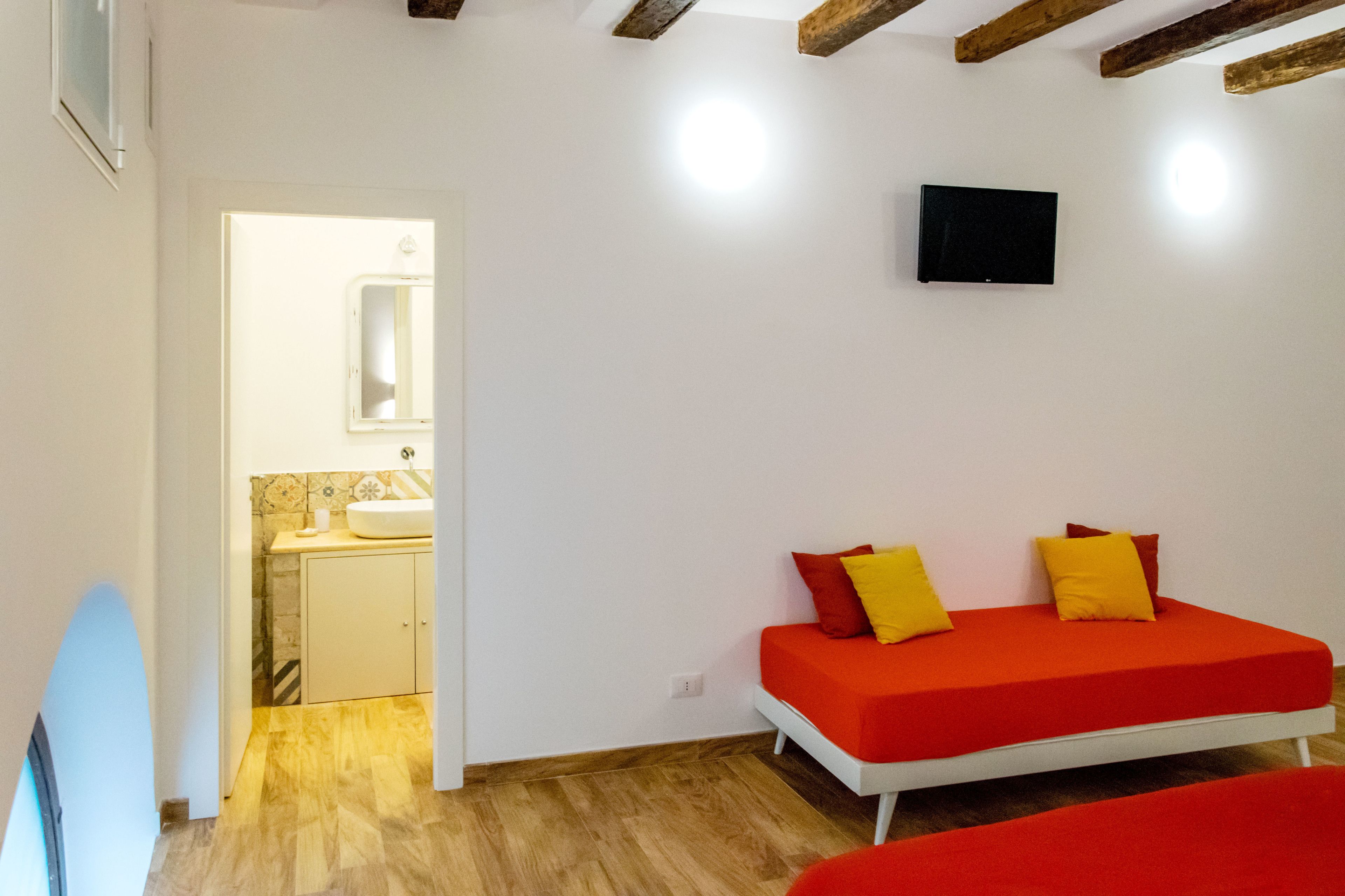 A room with a wooden floor, a red sofa with yellow cushions, a white wall, and a small TV mounted on the wall.
