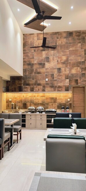 A modern dining area with a high ceiling, a ceiling fan, and a stone-tiled wall. There are dining tables and chairs, and a buffet counter with food items. - Zaith Residency, Chennai (Chennai)