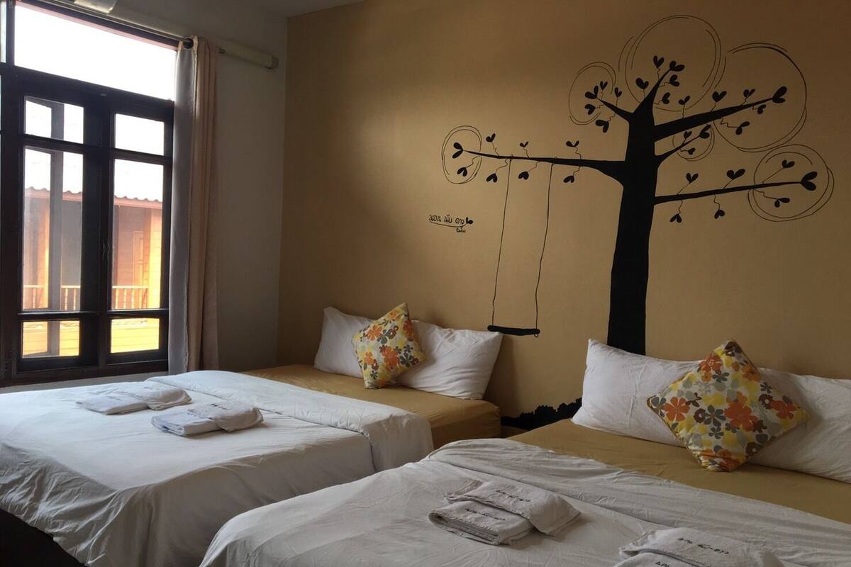 A hotel room with two beds, each with white linens and floral-patterned pillows. There is a large tree mural on the wall, and a window with curtains.
