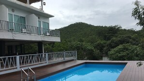 Una piscina con terraza de madera, un balcón con barandilla y vistas a una colina boscosa.