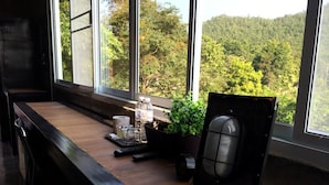 Un escritorio de madera con una computadora, una planta en maceta y un calentador en una habitación con ventanas amplias que dan a un bosque.