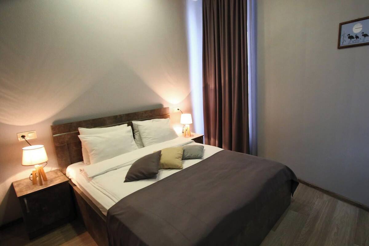 A hotel room with a neatly made bed, two bedside lamps, a wooden headboard, and a framed picture on the wall.