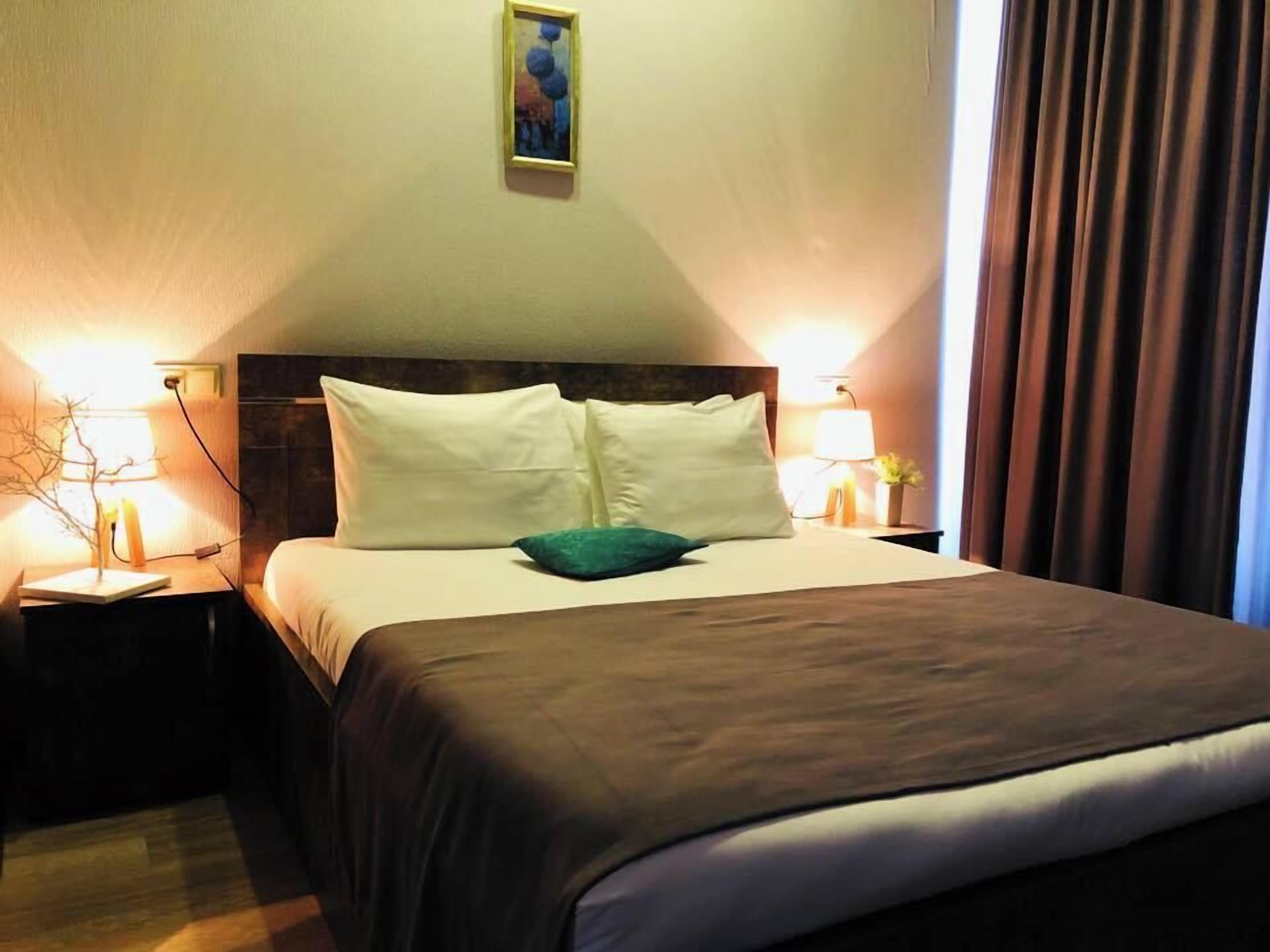 A hotel room with a large bed, two bedside tables with lamps, a painting on the wall, and a window with curtains.