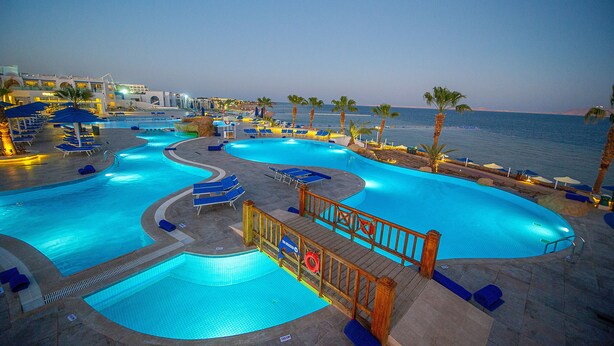 Une grande piscine extérieure divisée en plusieurs zones, entourée de chaises longues et de palmiers, avec une vue sur la mer en arrière-plan.