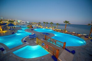 A large outdoor swimming pool with multiple sections, surrounded by lounge chairs and palm trees, with a view of the sea in the background.