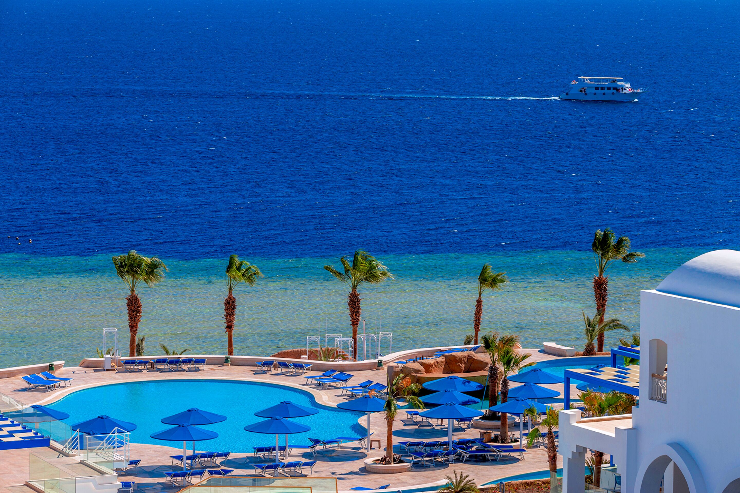 A resort pool with blue umbrellas and lounge chairs, surrounded by palm trees, with a view of the ocean and a boat in the distance.