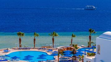 A resort pool with blue umbrellas and lounge chairs, surrounded by palm trees, with a view of the ocean and a boat in the distance.