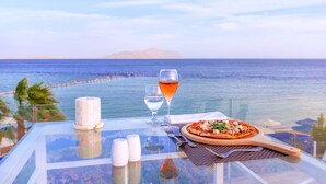 Une table avec une pizza, un verre de vin, des salières et des poivrières, et une bougie, donnant sur une plage avec l’océan et une montagne au loin.