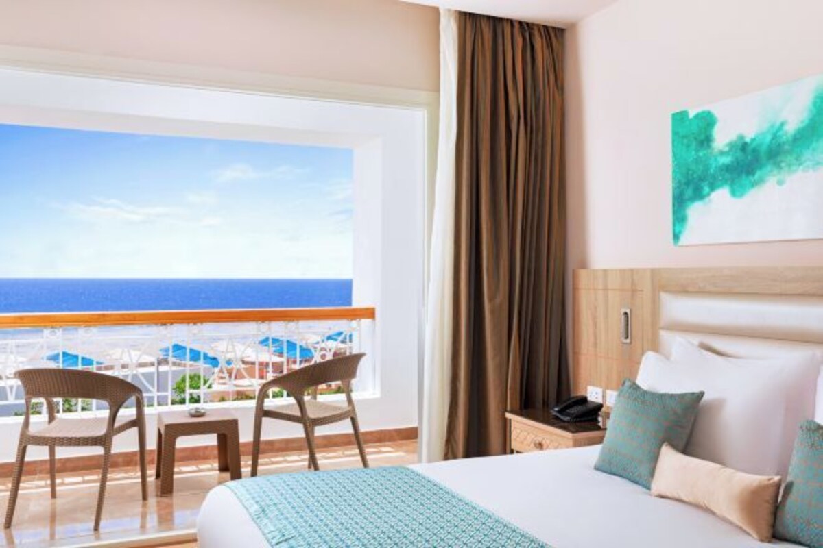 Een hotelkamer met een bed, twee stoelen, een nachtkastje, een schilderij en uitzicht op de oceaan.