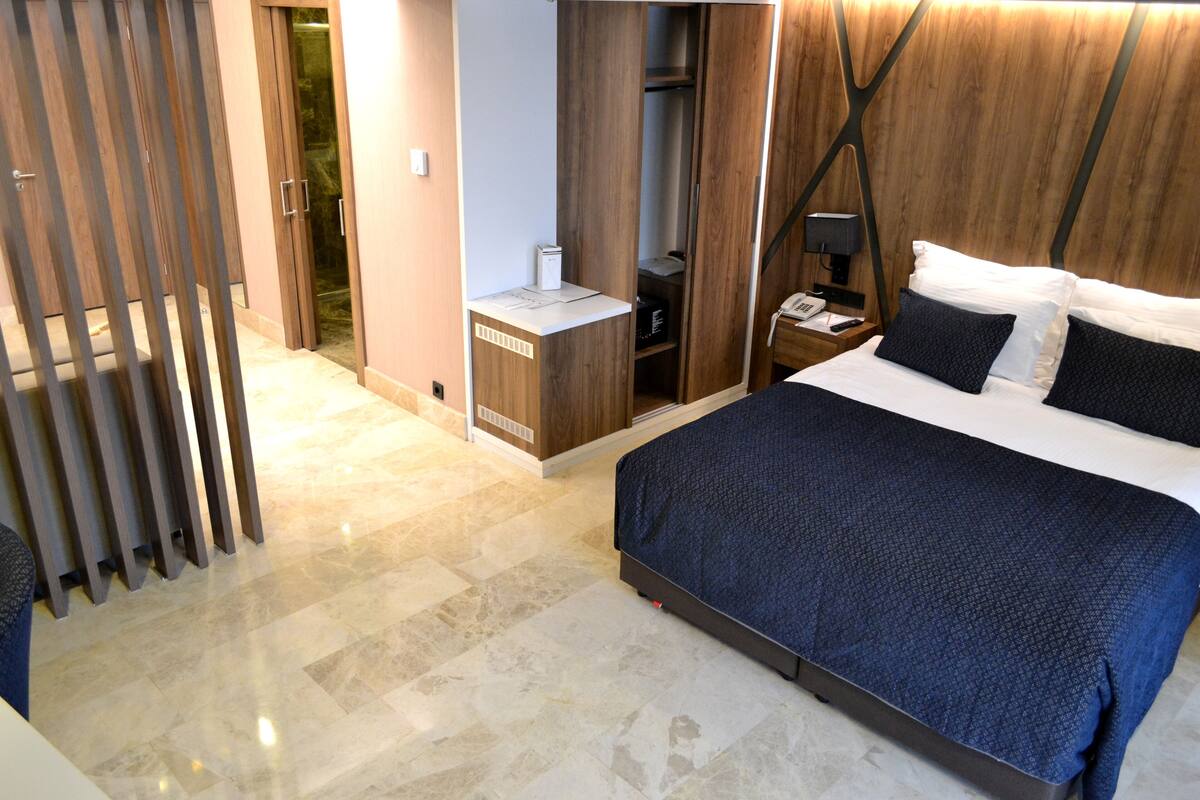 A modern hotel room with a large bed, a wooden wardrobe, a small desk, and a walk-in closet.