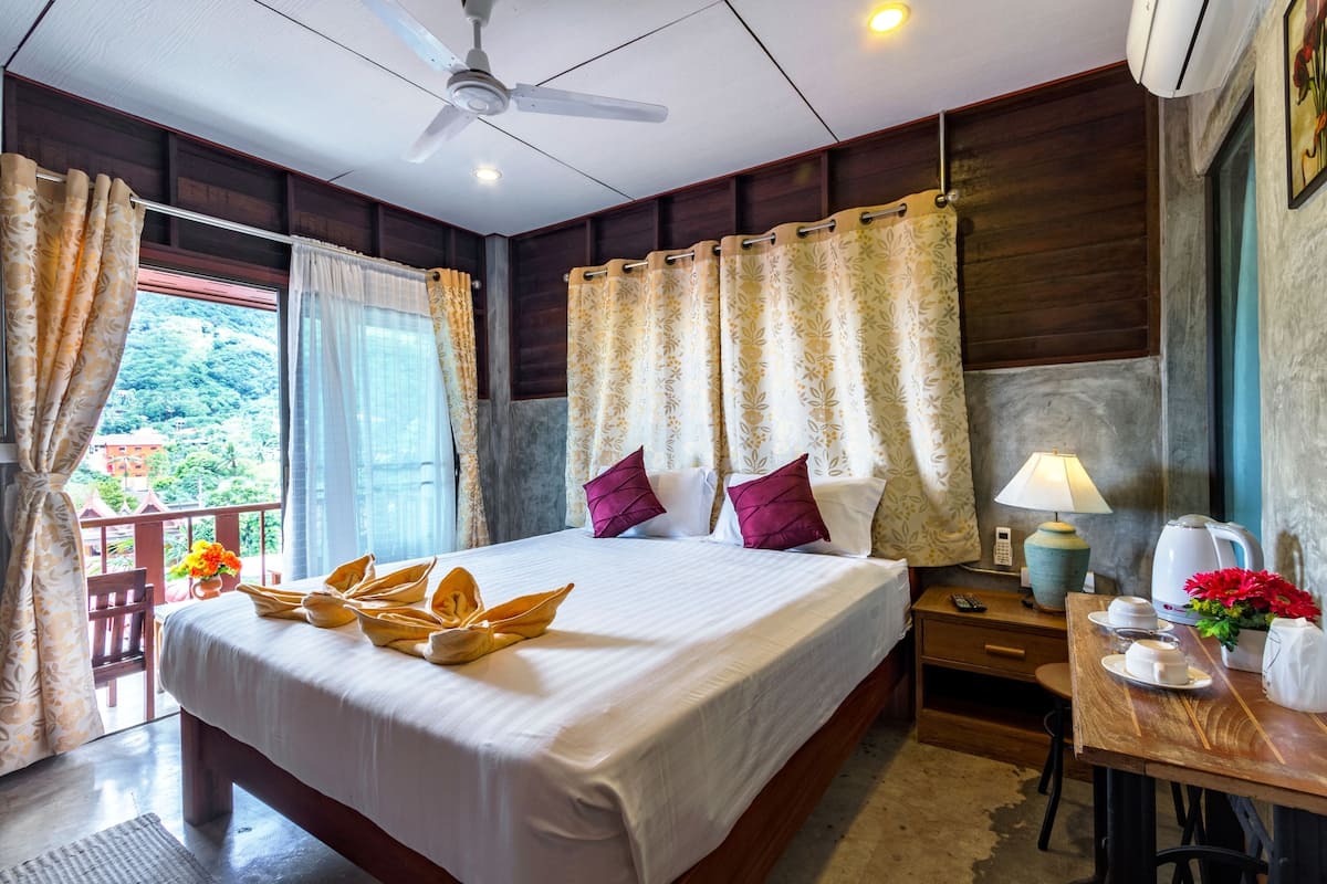 A hotel room with a bed, a desk, a chair, a table with a lamp, a coffee maker, and a view of greenery outside.