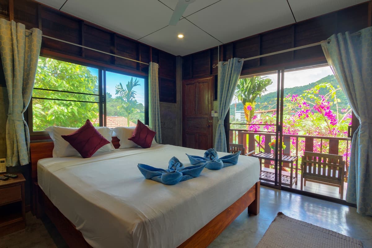 A large bed with white linens and blue towels, wooden headboard, and a view of lush greenery and flowers through a balcony.