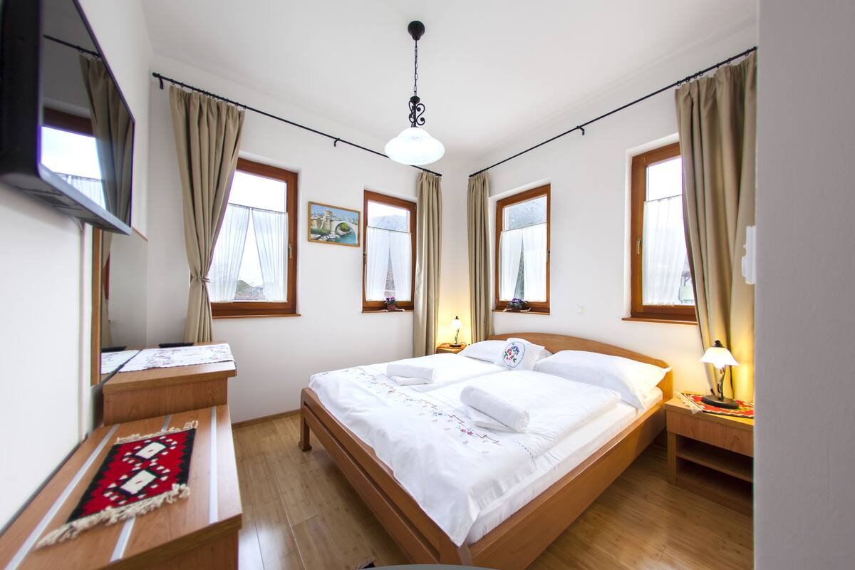 A hotel room with a bed, bedside tables, a TV, and windows with curtains.
