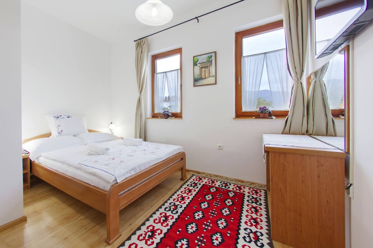 A bedroom with a bed, a wooden dresser, a window with curtains, a wall-mounted light fixture, and a rug.