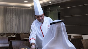 Un cuiner amb uniforme blanca i gorro de cuina preparant menjar a una taula en un restaurant.