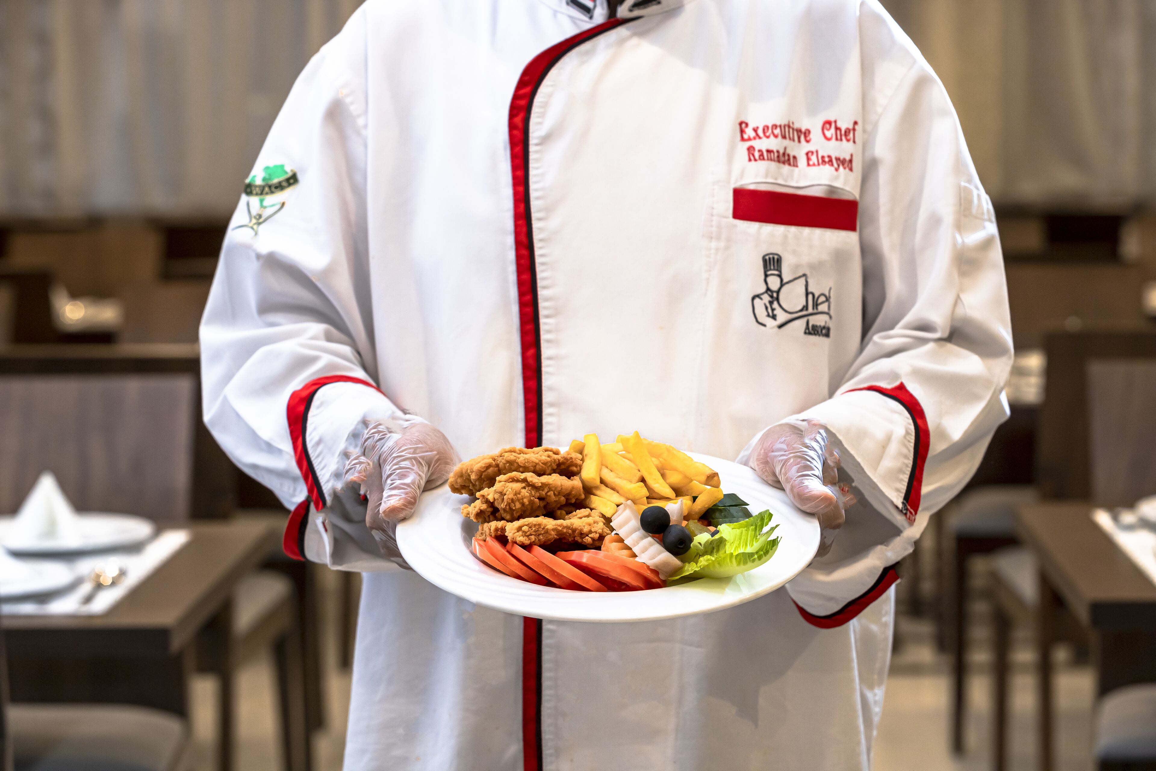 A person in a white chef's uniform holding a plate with fried chicken, fries, carrots, and olives.
