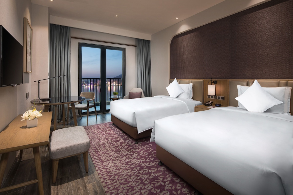 Une chambre d’hôtel avec deux lits, un bureau, une chaise, une télévision et un balcon donnant sur la ville.