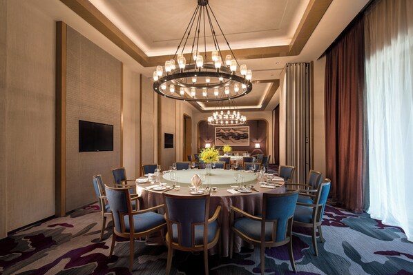 A hotel dining room with a round table set for a meal, surrounded by chairs, a chandelier, and a large painting on the wall.