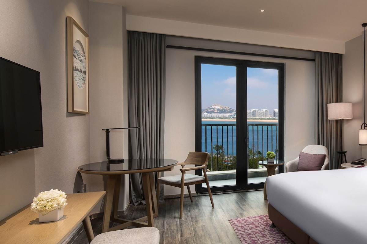 Une chambre d’hôtel équipée d’un lit, d’un bureau, d’une chaise, d’une télévision et d’un balcon offrant une vue sur la mer et les bâtiments.