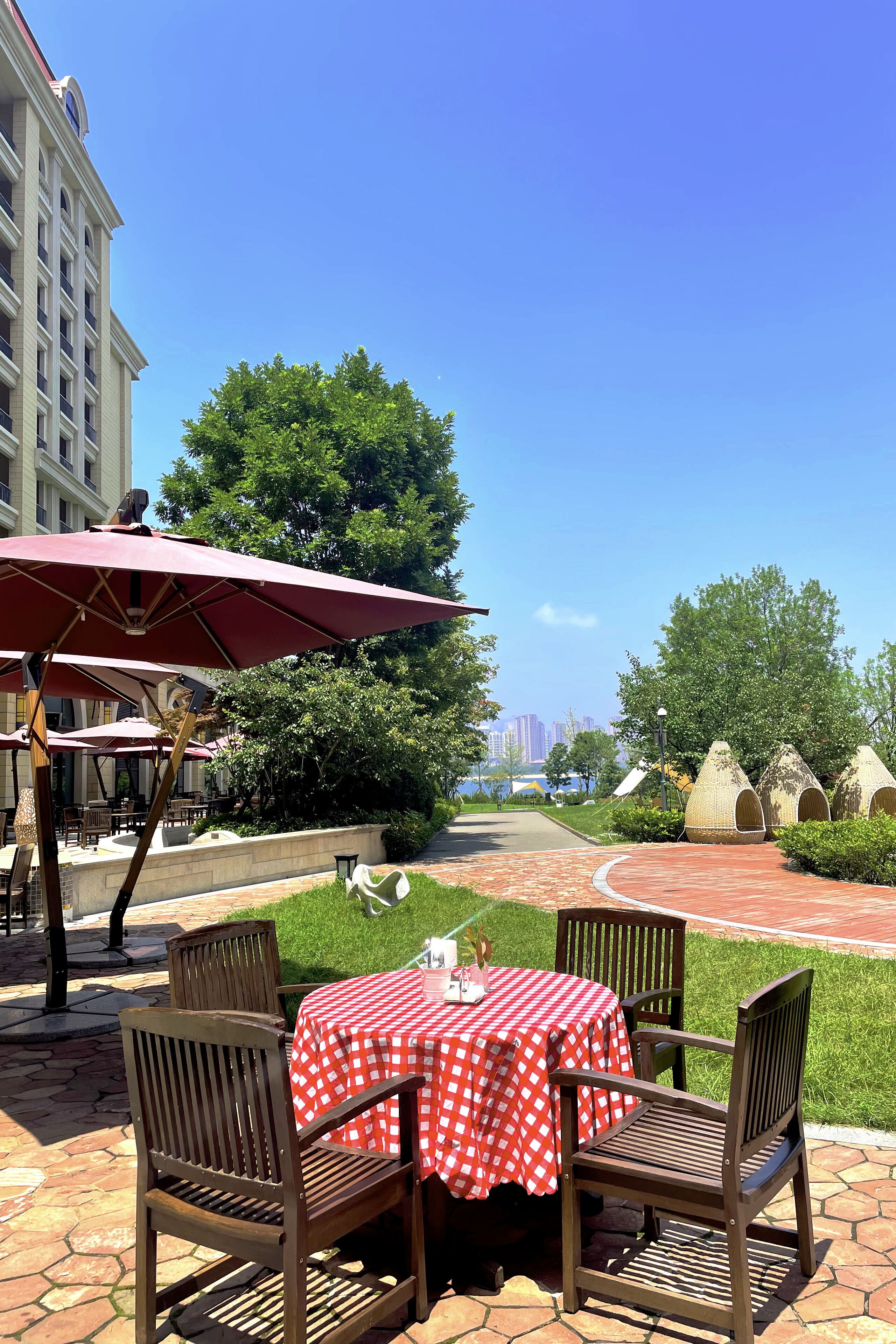 Een terras buiten met een rood-wit geruit tafelkleed, houten stoelen en een rode parasol.