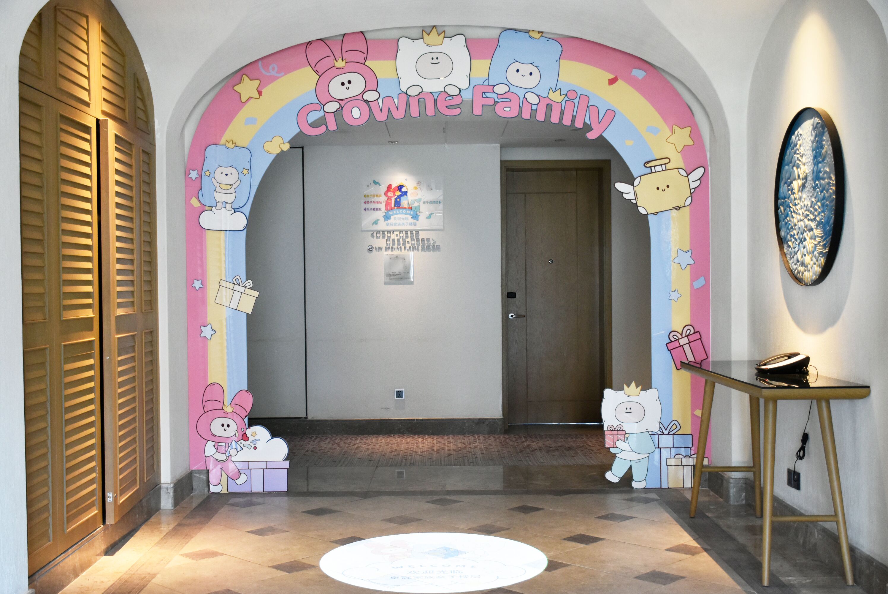 Um corredor com um arco colorido identificado como “Crown Family” e personagens de desenho animado. Há uma porta de madeira, uma pequena mesa e uma obra de arte na parede.
