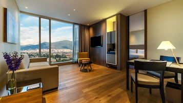 Habitación de hotel moderna con un ventanal grande que ofrece vistas a la ciudad, un sofá, una mesa de comedor con sillas, un televisor montado en la pared y una lámpara sobre la mesa.