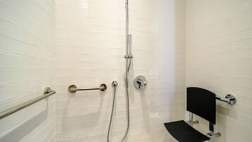 A bathroom with white tiled walls, a shower with a handheld hose, a grab bar, and a black bench.