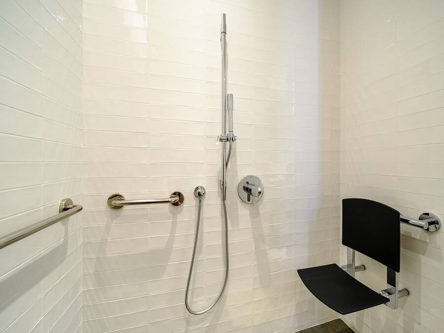A bathroom with white tiled walls, a shower with a handheld hose, a grab bar, and a black bench.