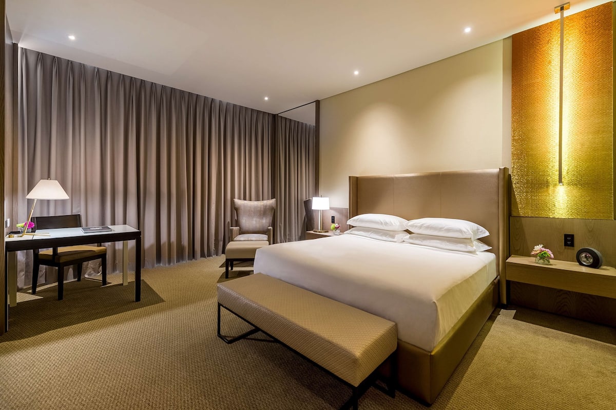 Ein Hotelzimmer mit einem großen Bett, einem Schreibtisch mit Stuhl, einer Bank, einem Nachttisch mit Lampe und einer Wand mit Strukturmuster.