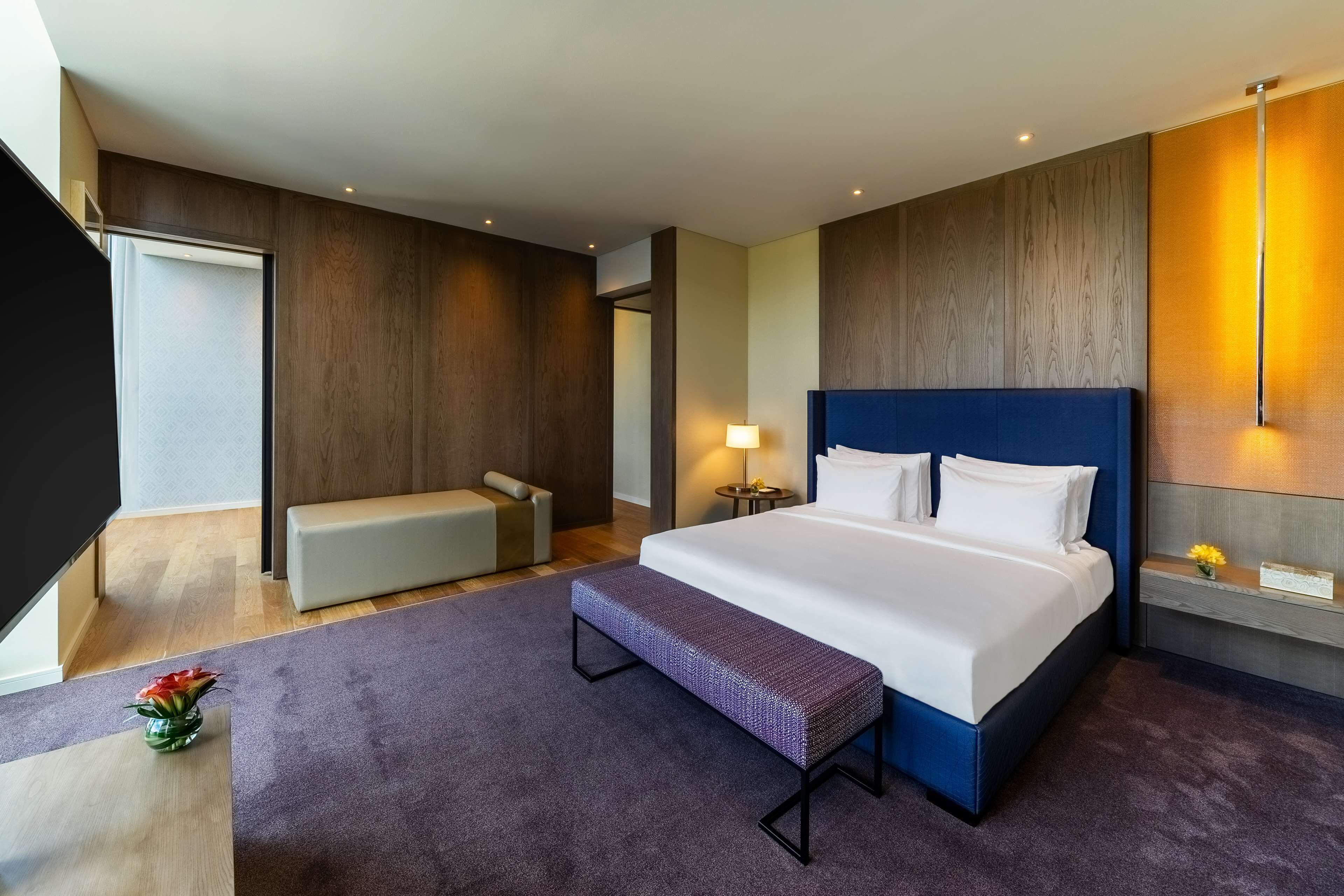 Una habitación de hotel moderna con una cama grande, un banco, una mesita de noche y una pequeña mesa con un florero.