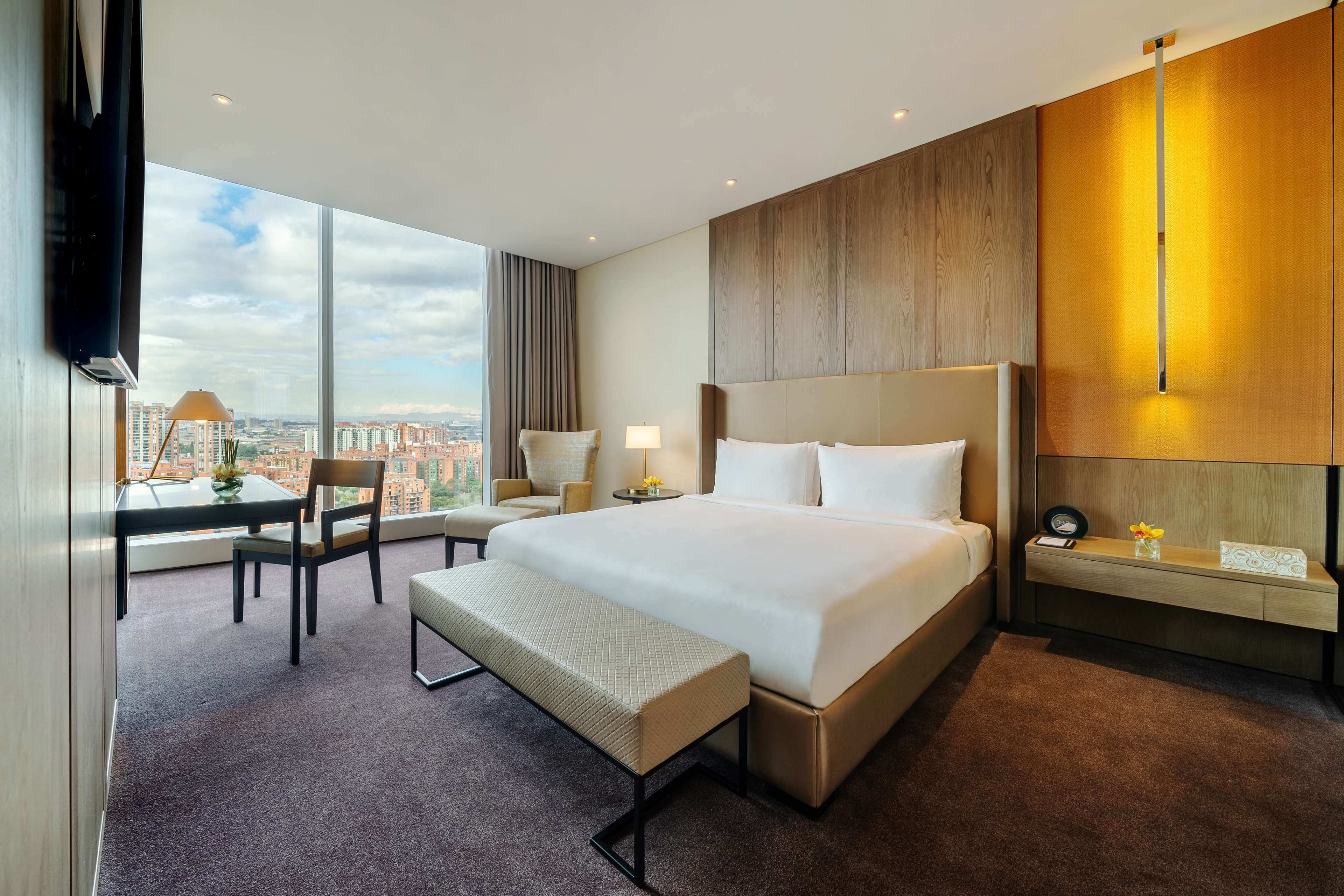 A hotel room with a large bed, a desk with a chair, a bench, a nightstand, and a view of the city.