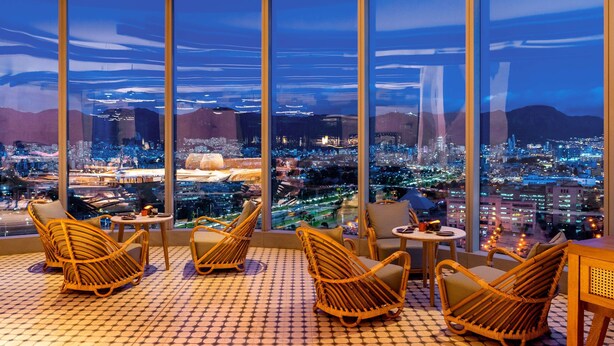 Ein Hotelzimmer in einem Hochhaus mit großen Fenstern und Blick auf die beleuchtete Stadt bei Nacht.