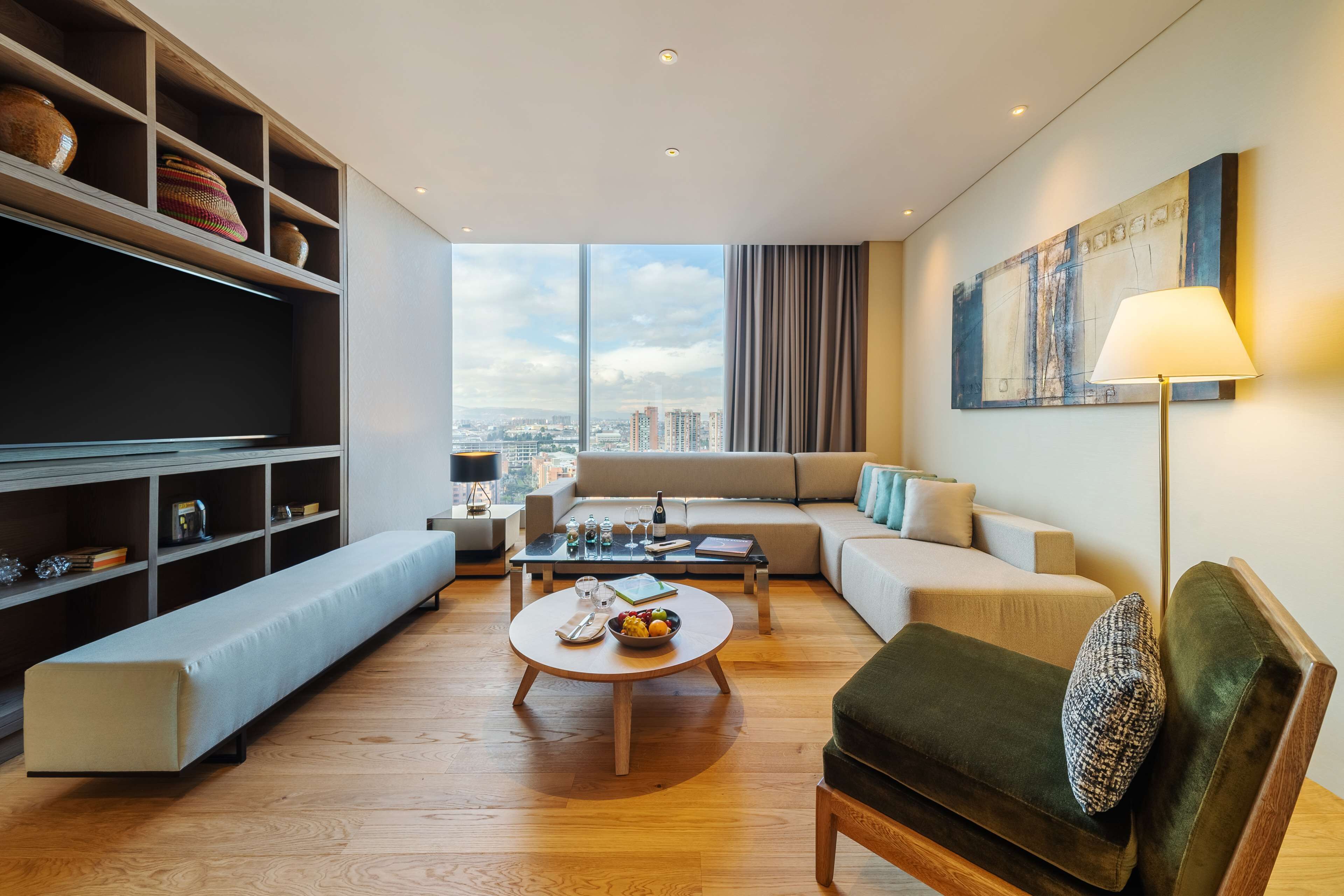 A modern living room with a sofa, armchair, and a coffee table with fruit. There is a bookshelf and a flat-screen TV.
