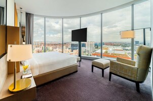 Habitación de hotel con una cama grande, un escritorio con lámpara, una silla y un televisor. La habitación tiene un ventanal con vistas a la ciudad.
