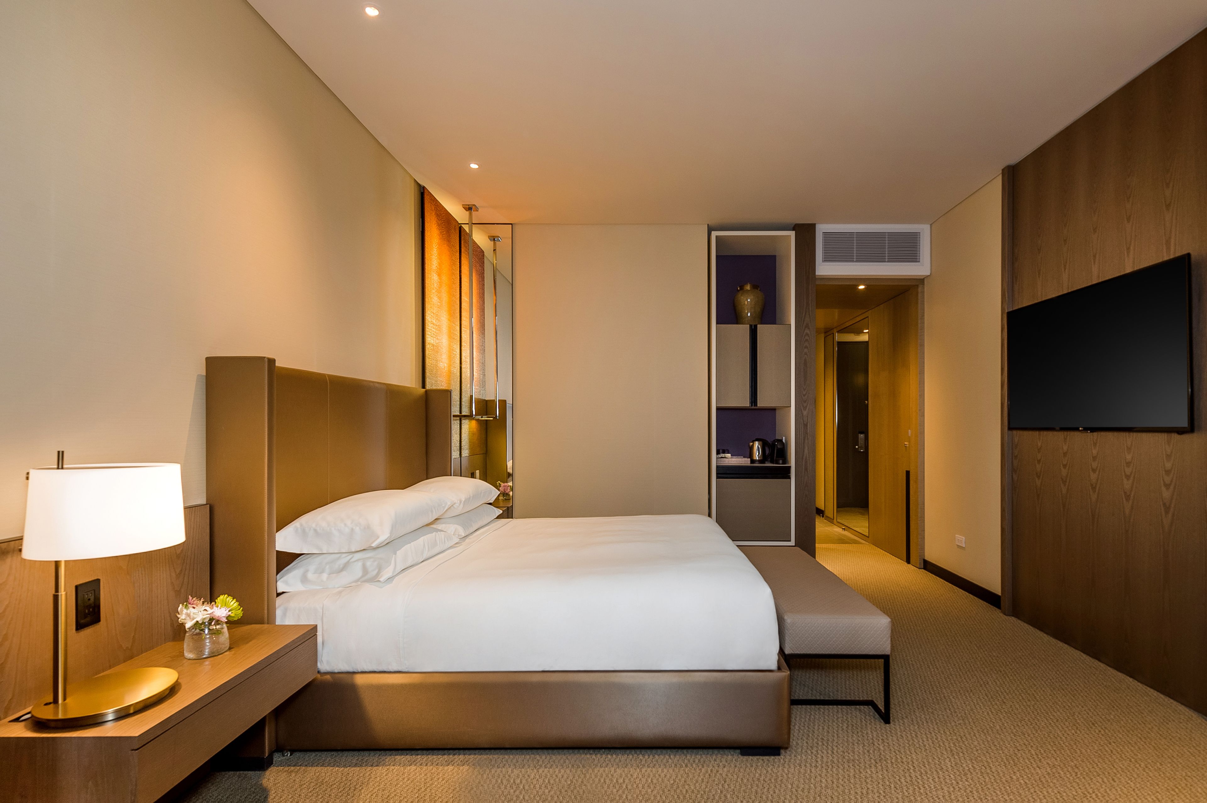 A hotel room with a large bed, bedside table, lamp, and a flat-screen TV mounted on the wall.