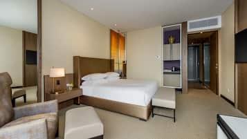 Habitación de hotel con una cama grande, un área de descanso con una silla y reposapiés, una mesita de noche con lámpara, un televisor montado en la pared y una pequeña cocina.