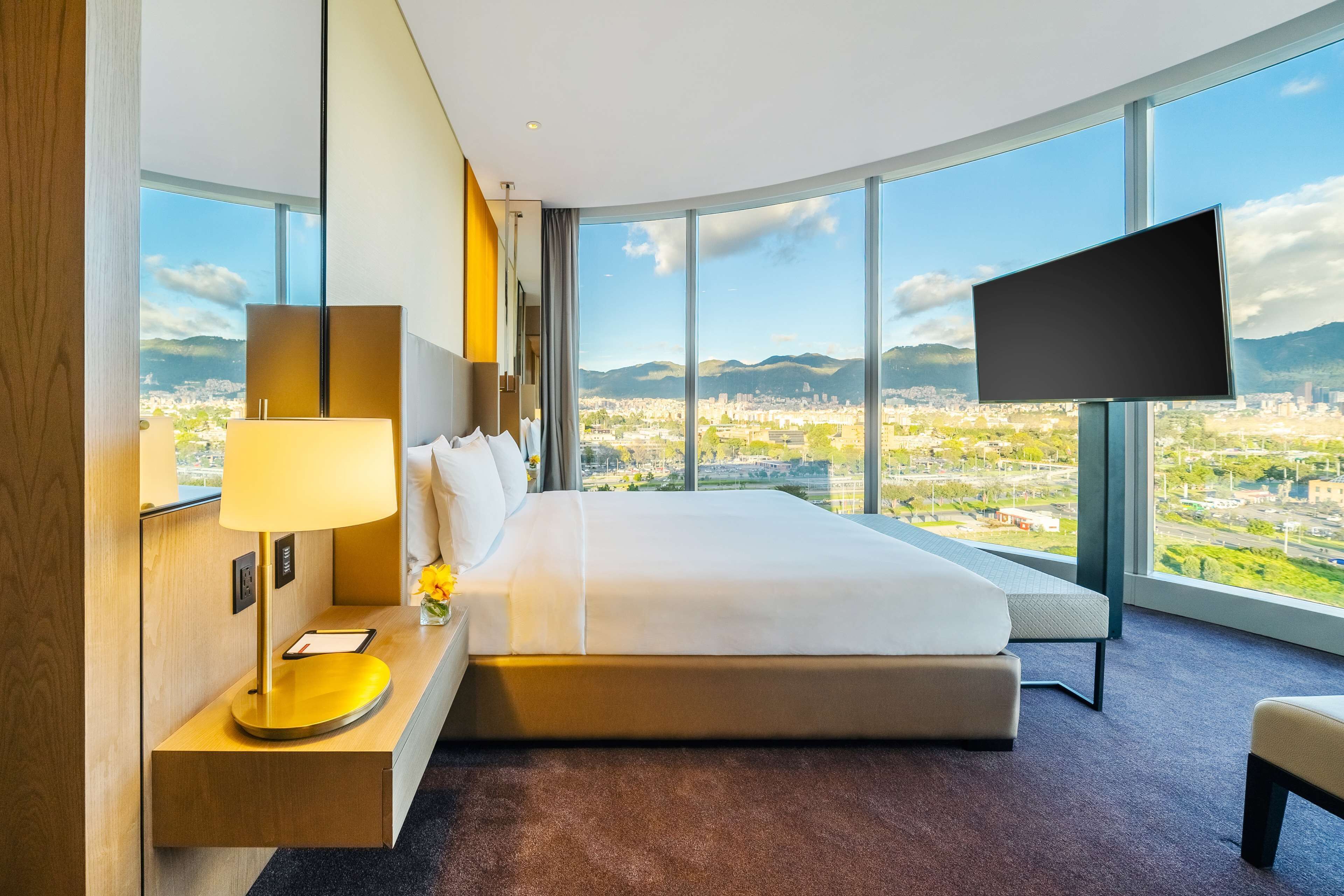 Ein modernes Hotelzimmer mit einem großen Bett, einem Flachbildfernseher und Blick auf die Stadt durch große Fenster.