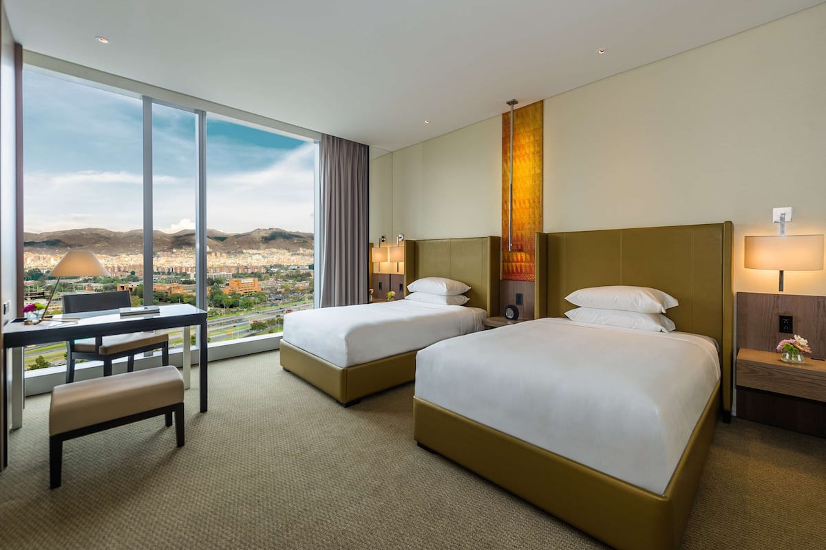 Ein Hotelzimmer mit zwei Betten, einem Schreibtisch und einem großen Fenster mit Blick auf die Stadt.