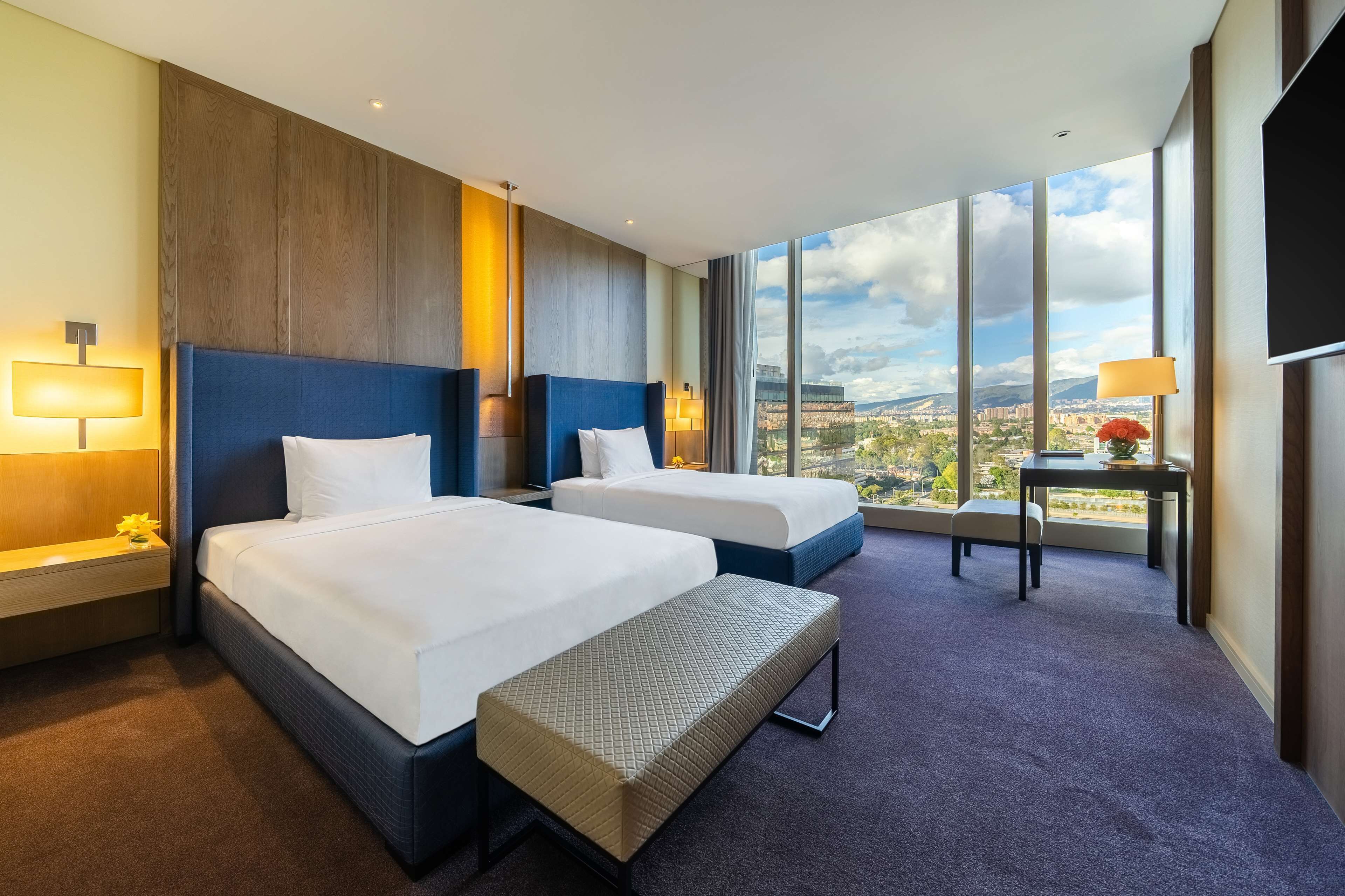 A hotel room with two beds, a large window with a city view, a small table with a lamp, and a bench.