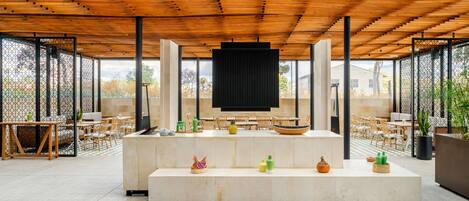 A spacious indoor area with a wooden ceiling, a central seating area with tables and chairs, and a large black screen.